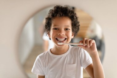Afrikalı Amerikalı genç bir çocuk bir aynanın önünde duruyor, elinde bir diş fırçası tutuyor ve dişlerini fırçalıyor. Görevine odaklanmış, tam bir temizlik sağlamak için yansımasına bakıyor..
