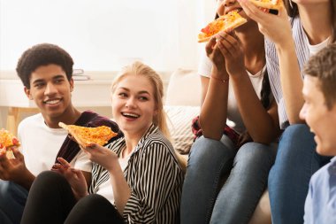 Neşeli bir grup genç arkadaş, iyi ışıklandırılmış bir oturma odasında, yerde ve kanepede rahat bir şekilde oturur, pizza dilimlerini paylaşır ve keyifli sohbetler ederler.