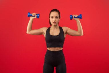 Bir kadın yüzünün önünde iki dambıl ile ayakta duruyor, antrenman yapıyor ya da egzersiz yapıyor gibi görünüyor. Ağırlıkları elinde tutarken yüzünde odaklanmış bir ifade var..