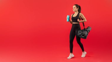 Siyah spor kıyafetleri giymiş formalı bir kadın yoga minderi ve spor çantası tutarken kendine güvenerek öne doğru adım atıyor. Beyaz spor ayakkabıları parlak kırmızı arka planla tezat oluşturuyor.