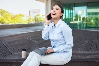 Asyalı bir kadın bankta oturuyor, telefonuyla konuşuyor, kucağında bilgisayarla. Odaklanmış görünüyor, muhtemelen önemli konuları tartışıyor.