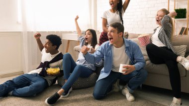Bir grup arkadaş sıcak bir oturma odasında toplanmış, neşe ve heyecan ifade ediyorlar. Bazıları kanepede otururken, diğerleri yerde oturup spor maçı veya TV 'de yarışma maçı izliyor..