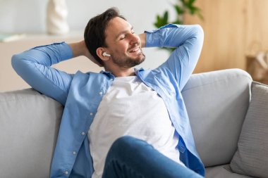 Gri bir kanepede rahat bir şekilde uzanan memnun bir adam elleri başının arkasında, gözleri kapalı, ve beyaz kablosuz kulaklıklarıyla müzik dinlerken mutlu bir gülümseme.