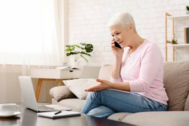 Son sınıf öğrencisi bir kadın, parlak bir oturma odasındaki krem rengi bir koltukta rahat bir şekilde oturuyor cep telefonuyla canlı bir konuşma yapıyor ve dizüstü bilgisayarının ekranına bakıyor.