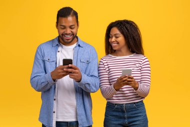 Neşeli genç bir Afro-Amerikalı erkek ve kadın yan yana durur, her biri kendi akıllı telefonuna dalmıştır. Gülümsemeler yüzlerini süsledikçe, neşeli bir anı paylaşıyor gibi görünüyorlar.