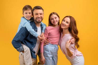 Bir baba, anne, oğul ve kızdan oluşan mutlu bir aile, neşeli ifadelerle birlikte yakın poz veriyor. Baba oğlunu sırtında taşır.