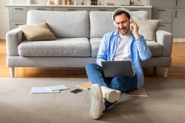 Bir adam yerde oturuyor, kulaklık takıyor ve dizüstü bilgisayar kullanırken dikkatlice müzik dinliyor. Dijital aktivitelerine odaklanmış ve meşgul görünüyor..