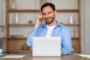 Bir adam dizüstü bilgisayarın önünde oturuyor, telefonla meşgul. Bilgisayarında çalışırken telefonla konuşurken odaklanmış gibi görünüyor..