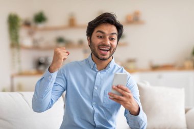 Hintli adam modern bir koltukta oturuyor, akıllı telefonunu elinde tutuyor. Cihaz ekranına odaklanmış görünüyor, muhtemelen mesaj ya da tarama yapıyor. Oda nötr renk düzeniyle iyi aydınlatılmış..