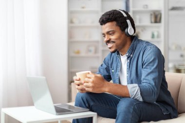 Neşeli, Afro-Amerikan bir adam beyaz bir koltukta rahat bir şekilde oturuyor, bir çift kulaklık takıyor ve elinde bir bardak tutuyor, müzikten zevk alıyor veya sanal bir sohbete dalıyor.