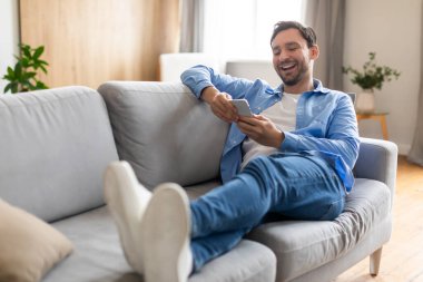 Bir adam rahat bir şekilde bir kanepede oturuyor, telefonuna dalmış. Elinde tuttuğu dijital cihaza kendini tamamen kaptırmış bir şekilde kaydırıyor ve yazıyor..