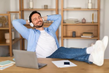 Bir adam masasında oturuyor, kulaklık takıyor ve dikkatle müzik dinliyor. Ses kaydıyla uğraşırken ifadesi odaklanmış durumda..