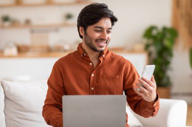 Arap adam bir kanepede oturuyor, aynı anda bir dizüstü bilgisayar ve cep telefonu kullanıyor. Kanepede rahat bir duruşla, cihazlarına odaklanmış görünüyor..