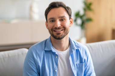 Neşeli bir yüzü olan bir adam koltuğa oturmuş, parlak bir gülümseme sergiliyor. Mobilyaların üzerinde otururken rahat ve mutlu görünüyor..