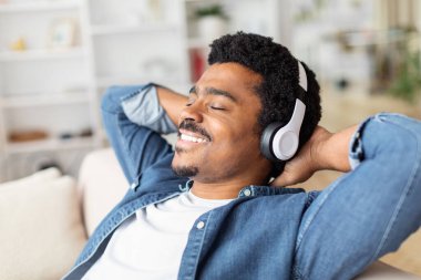 Afro-Amerikalı bir adam kanepeye oturmuş, kulaklık takıyor. O, koltukta dinlenirken kulaklıkla müzik veya ses içeriği dinlemeye odaklanmış gibi görünüyor..