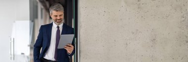 Ofis binası koridorunda dikilirken dijital tablet kullanan yakışıklı orta yaşlı işadamı, takım elbiseli gülümseyen erkek girişimci, modern bir cihaz, iş uygulaması, kopyalama alanı