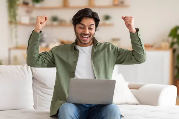 stock image Arab man is seated on a couch, with his arms raised while focused on using a laptop in front of him. He appears engaged and concentrated on the screen, possibly working or browsing the internet.