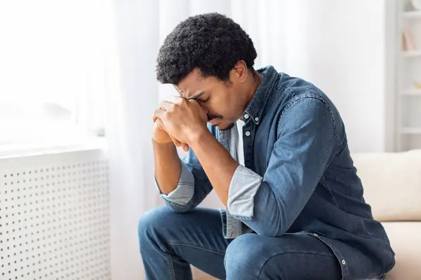 stock image Black man is seated on a couch, with one hand pressed against his face in a contemplative gesture. He appears deep in thought, possibly experiencing emotions like stress, sadness, or exhaustion.