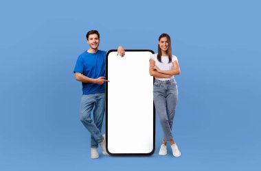 Yanyana duran neşeli genç bir çift, sağlam mavi bir arka planda büyük, boş bir cep telefonu ekranı sunuyorlar..