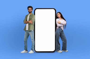 Neşeli genç bir Hintli erkek ve kadın büyük, boş ekranlı bir akıllı telefonun yanında dururlar, kolları çapraz bir şekilde seyirciye çarpıcı mavi bir arka plana karşı dururlar.