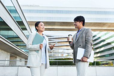 İki iş adamı, takım elbiseli Asyalı bir adam ve iş elbiseli bir kadın modern bir ofis binasının önünde sohbet ediyorlar. Muhtemelen işle ilgili konuları tartışıyorlar.