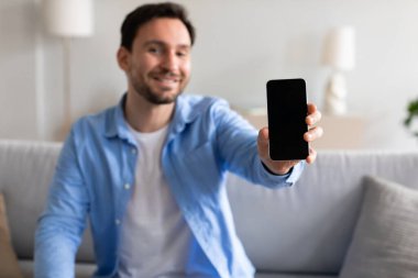 Mavi gömlekli neşeli bir adam kollarını uzatıyor ve boş ekranlı bir akıllı telefonu gururla gösteriyor. Gün boyunca güzel ışıklandırılmış, rahat bir oturma odasında bir kanepede oturuyor..