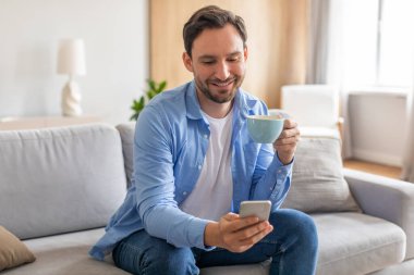 Bir adam kanepede oturuyor, bir elinde bir fincan kahve, diğer elinde telefonuna bakıyor. Rahatlamış ve telefon ekranına odaklanmış görünüyor..