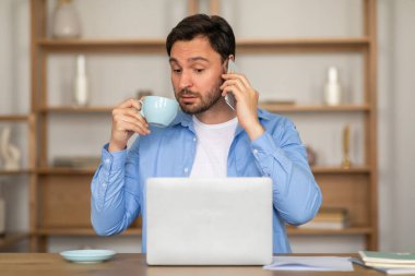 Bir adam elinde bir fincan kahveyle ayakta duruyor, telefonuyla sohbet ediyor. Telefonu kulağına dayadığı anda telefona odaklanmış gibi görünüyor..