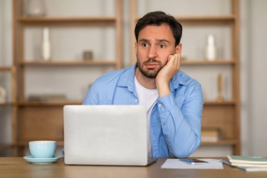 Genç bir adam, iyi ışıklandırılmış bir odada bir masada oturuyor, bir dizüstü bilgisayar ekranına boş boş bakarken yanağında dinlenirken can sıkıntısı veya yorgunluk belirtileri gösteriyor.