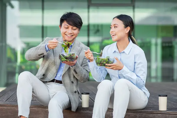 stock image Two Asian individuals, dressed in business attire, are seen seated at a table. Both are actively engaged in consuming a bowl of salad, showcasing a healthy meal choice during their work break.