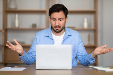 Sıradan mavi gömlekli bir adam masanın üzerinde oturuyor ve dizüstü bilgisayarına bakarken belirsizlik içinde kollarını kaldırmış şaşkın bir yüz ifadesi sergiliyor, şaşırtıcı bir iş durumuyla karşı karşıya.