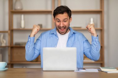 Bir adam elinde dizüstü bilgisayarla masanın önünde kutlama yapıyor. Heyecanlı ve neşeli görünüyor, muhtemelen bir amaca ulaşıyor ya da iyi haberler alıyor..