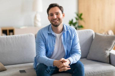 Oturma odasındaki bej bir kanepede oturan bir adam. Odada bir sehpa, bir lamba ve bir kitaplık bulunuyor. Adam rahat görünüyor ve telefonuna bakıyor..