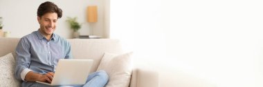 Evdeki kanepede oturan mutlu genç adam, bilgisayar laptopu kullanıyor, web sörfü yapıyor ya da internette çalışıyor, kopyalama alanı olan panorama