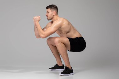 Gri arka planda bir adam çömelme egzersizi yaparken, dizlerini bükerken ve belini düz tutarken görülüyor. Alt vücut kasları için bir güç antrenmanı yapıyor..