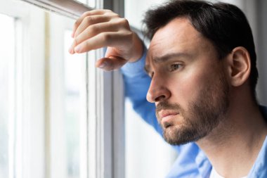 Bir adam pencerenin kenarında durup, düşünceli bir ifadeyle dışarı bakıyor. Doğal ışık, dışarıdaki manzarayı izlerken profilini ön plana çıkarıyor..