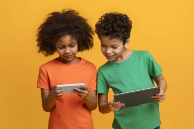 İki Afrikalı Amerikalı genç çocuk, bir oğlan ve bir kız, yerde birlikte oturuyorlar. Her ikisi de ekrana takılmış bir tablet bilgisayar kullanıyorlar. Arka plan parlak sarı bir renktir.