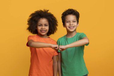 İki neşeli Afrikalı Amerikalı genç kardeş canlı sarı bir arkaplanda yan yana duruyorlar. Parlak, koyu renkli tişörtler giyiyorlar ve yumruklarını sımsıkı birleştirirken gülümsüyorlar.