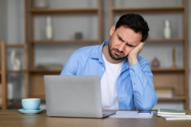 Genç bir adam ahşap bir masada oturuyor, önünde kapalı bir dizüstü bilgisayarla yanağına yaslanırken sıkıntılı görünüyor. İfadesi zorlu bir sorunla baş etmeyi öneriyor.