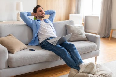 Orta yaşlı bir adam rahat bir kanepede rahatlarken görülüyor, dizüstü bilgisayarına sarılmış bir şekilde. Çalışırken sakin ve odaklanmış görünüyor ya da kapalı bir ortamda internette geziniyor..