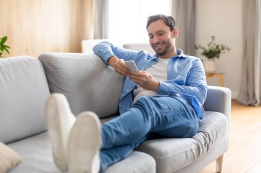 Mutlu adam, akıllı telefonuyla ekranda odaklanmış bir şekilde koltuğa yaslanıyor. Duruşu rahat, bu da içeride rahat bir ortam olduğunu gösteriyor..
