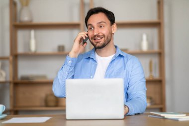 Masada oturan, telefonla konuşan bir adam. Önünde bir dizüstü bilgisayarla birden fazla iş yapıyor..