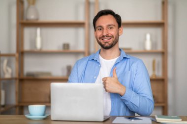 Bir adam bir masada oturuyor, dizüstü bilgisayar kullanıyor. Gülümsüyor ve baş parmak işareti yapıyor, işindeki başarısını ya da onayını gösteriyor..