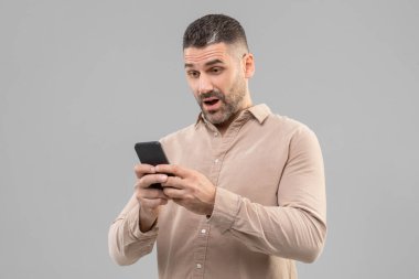 Kısa saçlı, elinde akıllı telefon tutan şaşırmış bir adam. Ekrana bakıyor. Cihazı kullanmaya odaklanmış gibi görünüyor..