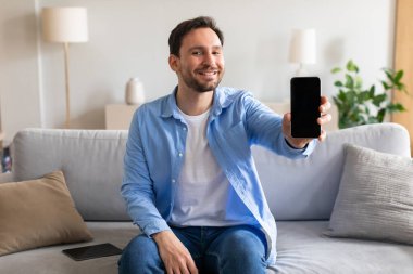 Kanepede oturan bir adam elinde bir akıllı telefon tutuyor. Ekrana odaklanmış görünüyor, muhtemelen bakınıyor ya da mesajlaşıyor. Oda rahat, yumuşak bir ışık sahneyi aydınlatıyor..