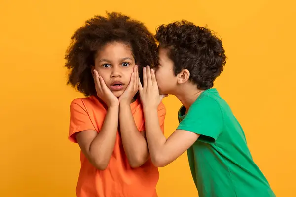 stock image A young African American boy in a green shirt is whispering into the ear of his surprised sister, who is wearing an orange shirt. Her hands are placed on her cheeks, and her expression is astonishment
