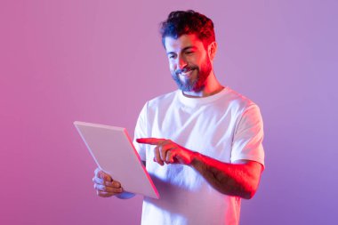 Beyaz tişört giyen sakallı bir adam, mor ve pembe neon ışıklı bir stüdyoda dikilirken dijital tabletle etkileşime giriyor. Gülümsüyor ve tabletteki içerikle meşgul görünüyor..