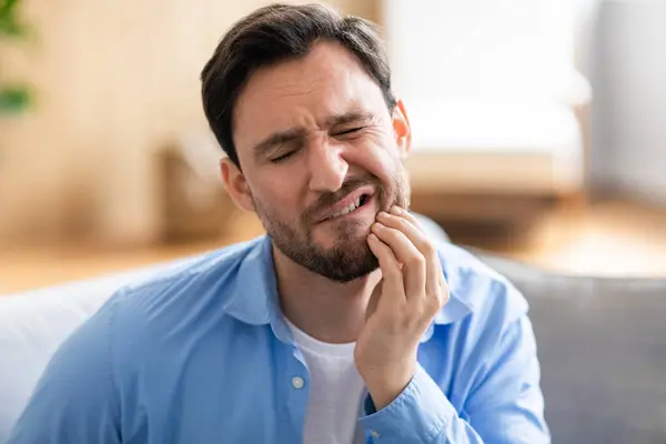 stock image A man is shown sitting on a couch, visibly in discomfort, holding his jaw with a pained expression due to a toothache. He appears to be seeking relief or contemplating seeking dental help.