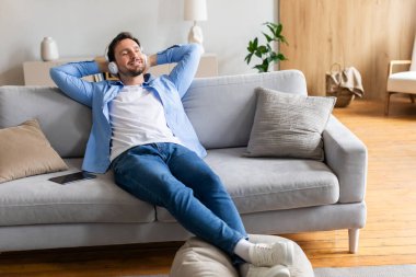 Oturma odasındaki kanepede rahatça oturan bir adam rahat ve rahat görünüyor. Oda doğal ışıkla aydınlanmış ve adam dinlenme ve dinlenme anının tadını çıkarıyor gibi görünüyor..