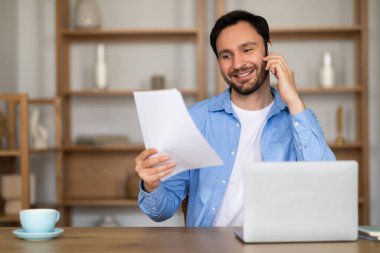 Bir adam telefonda konuşurken masada oturuyor, elinde bir kağıt parçası tutuyordu. Telefonla iletişim kurarken odaklanmış görünüyor ve aralıklı olarak gazeteye bakıyor..